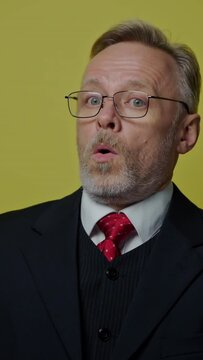 Positive businessman showing different emotions. Mature man in glasses and elegant costume shows funny face on the camera on yellow studio background. Vertical video
