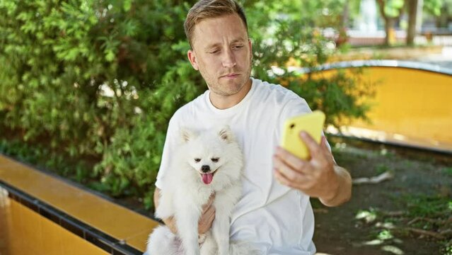 Cheerful young caucasian man sitting with his happy dog on park bench, enjoying the outdoors while confidently making a funny selfie picture on his smartphone