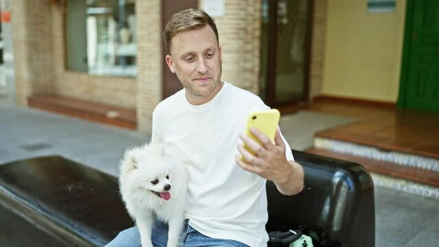 Cheerful, young caucasian man sitting on city street bench, joyfully taking a confident, happy selfie with his handsome pet dog using smartphone.