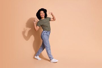 Fototapeta na wymiar Full length photo of cheerful woman wear khaki t-shirt jeans trousers show v-sign go empty space isolated on beige color background