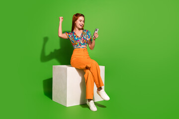 Full size photo of pretty young woman sit cube raise fists hold phone wear top isolated on bright...