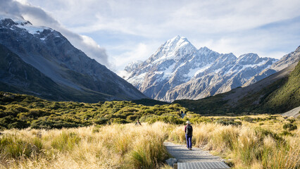 Tourists walking the trail in beautiful alpine valley with huge snowy mountain, Mt Cook, New Zealand - obrazy, fototapety, plakaty