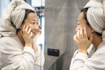 Portrait of beautiful Asian girl in bathrobe and with towel on her head cleaning her face and...