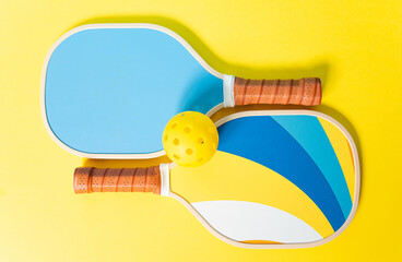 Photograph of two pickleball rackets and a ball on a yellow background. The rackets are blue and...