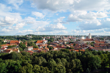 Fototapeta na wymiar Top view from the mountain of the architecture of the old city of Vilnius with old buildings with orange tiled roofs, town halls, churches, towers, cathedrals, green deciduous forest, sunlight, clouds