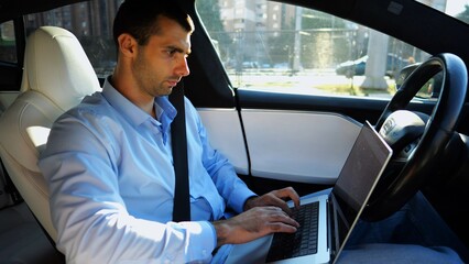 Handsome entrepreneur working on notebook while riding an autonomous self driving electric car at urban road. Male businessman typing text on laptop during riding on electrical vehicle with autopilot - obrazy, fototapety, plakaty