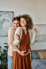 Happy couple expecting baby, man hugging his pregnant wife from behind with cheerful smile