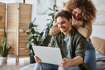 Sitting together with their laptop, curly young woman and brunette man enjoying a peaceful morning