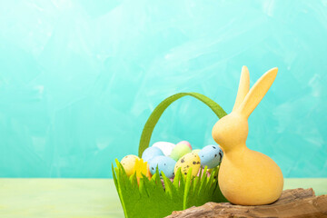 Yellow Easter bunny and green felt basket with decorative colorful eggs on turquoise. Copy space.