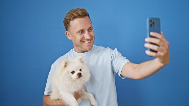 Cheerful young caucasian man taking a fun selfie with his happy pet dog on a smartphone, standing against an isolated blue wall background, expressing pure joy and confidence.