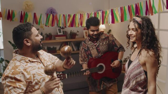 Full shot of three young cheerful Mexican people having fun at house party while celebrating national holiday - men in bright shirts playing guitar and maracas, woman in dress, scarf on hair dancing
