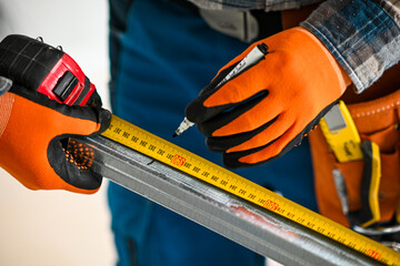 a construction worker in overalls, gloves and a tool belt measures the length of a plasterboard profile with a meter - close-up view - obrazy, fototapety, plakaty