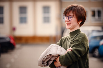 A cheerful, young woman in stylish eyewear and a cozy green sweater holds a jacket, showcasing a...