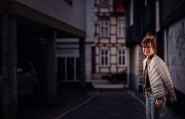 A fashion-forward woman with glasses and a green sweater carries her jacket confidently in a dimly lit urban alley.