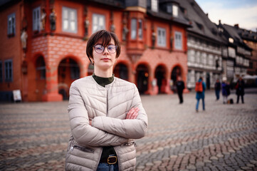 A woman stands confidently in a historic town square, her arms crossed, with traditional architecture creating a charming backdrop.