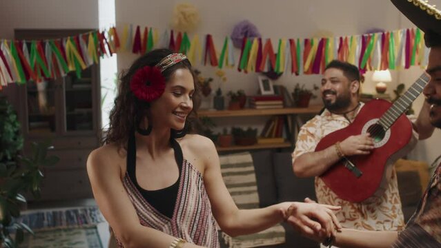 Medium footage of young Mexican couple, guy in sombrero hat, curly girl wearing dress, flower in hair, dancing together, while male friend is playing guitar and singing, celebrating national holiday
