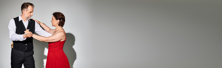 Portrait of mature attractive couple in red dress and suit dancing on grey background, banner