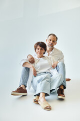 Middle aged couple wearing white shirts and jeans sitting on grey background and looking at camera