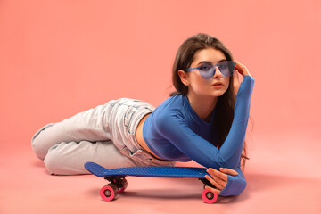 A young beautiful girl in a blue jacket and denim pants with a mini skateboard on a pink background.