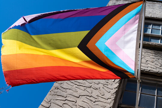  Old A LGBTQ+ rainbow progress pride flag flaps from a flagpole in a brisk breeze. Blue sky and trees behind in |New York, USA
