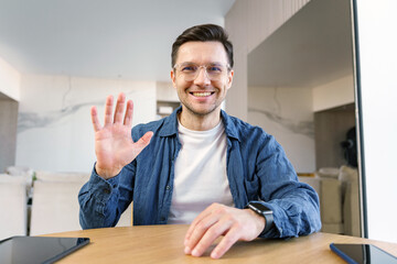 A genial man waves at the camera, with a tablet and laptop on the table, in a modern home environment. - obrazy, fototapety, plakaty