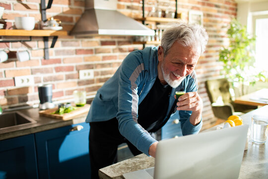 Senior man laughing while drinking juice and using laptop in kitchen