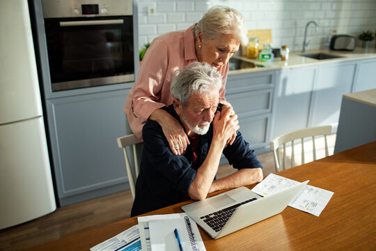 Concerned senior woman comforting man with laptop and bills at kitchen table