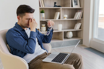 A man appears stressed or fatigued, taking a moment while working on his laptop in a serene home...