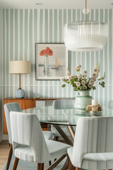 A dining room with striped wallpaper of light blue and white stripes, a modern glass table and chairs covered in linen fabric, a wooden sideboard, wall art hanging on the walls, a hanging lamp.