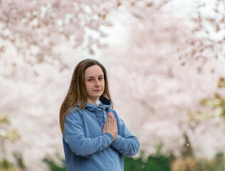 A young woman is engaged in breathing yoga practice against the backdrop of a spring blossoming sakura alley. Hands folded in a namaste gesture. Concept of mental, physical health and balance.