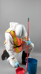 an unidentified person likley a man in a hazmat suit with rubber gloves and rubber boots with a hard hat and safety vest cleaning the floor with a broom and dustpan isolated on grey