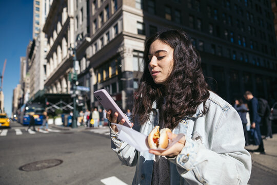 Woman eating a hot dog in New York and using smartphone