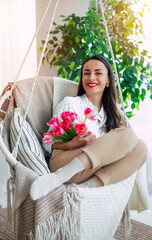 Vertical image of cute smiling brunette woman with bunch of flowers in hands is relaxing and swinging on hanging chair. Present in women's day from beloved man. St. Valentines day - 771004213