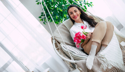 Beautiful smiling tender brunette woman with bunch of flowers is relaxing and swinging on hanging chair. Present in womens day from beloved man. St. Valentines day - 771004232
