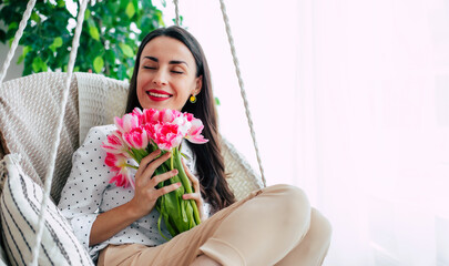 Attractive calm young female with closed eyes is smells the aroma of flowers while swinging on hanging chair at home. Gift in women's day with love. St. Valentines day, March 8, Birthday - 771004273