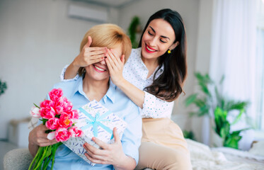 Mother and daughter. Attractive brunette stylish woman covering her mother's eyes while giving her present and gives a bouquet of flowers to tulips. Mothers day, birthday or womens day concept - 771004403