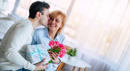 Side view photo of happy handsome man while he greeting and kissing his smiling mother with some holiday in domestic room. Mothers day, birthday or womens day concept - 771004410