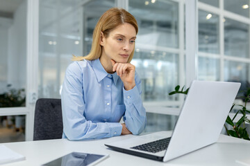 Fototapeta premium Professional female executive analyzing data on laptop while sitting at a white desk in a bright, contemporary office space.