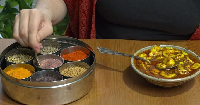 Homemade garlic pickle in a bowl and Indian spices box or masala box with all spices used to make garlic pickle. A woman scoops pickles with a spoon.