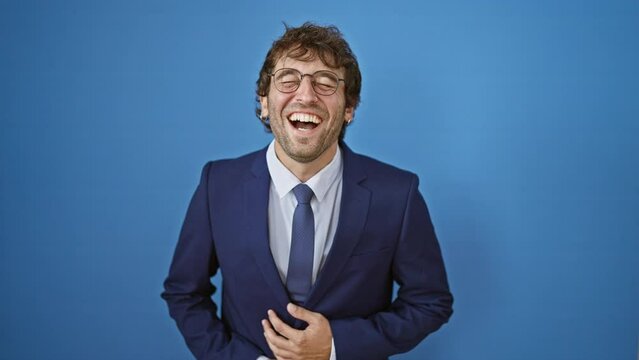 Joyful young man in a business suit laughs out loud, flashing a bright, handsome smile and clasping his hands in sheer delight! isolated against blue background.