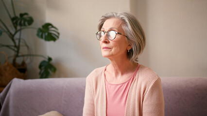 Portrait of beautiful mature old 60s woman, older middle aged female customer watching at camera and smiling while sitting on couch at home