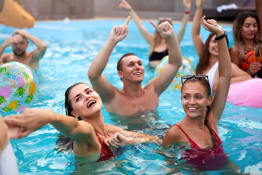 Friends have night pool party in a private villa swimming pool. Happy young people in swimwear splashing water, dancing with floaties and inflatable mattress, ball in a luxury resort.