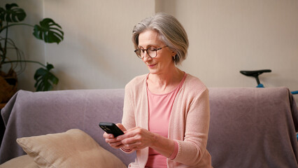 Mature old 60s woman, older middle aged female customer holding smartphone using mobile app, texting message, search ecommerce offers on cell phone technology device sitting on couch at home