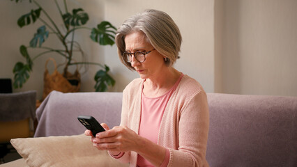 Mature old 60s woman, older middle aged female customer holding smartphone using mobile app, texting message, search ecommerce offers on cell phone technology device sitting on couch at home