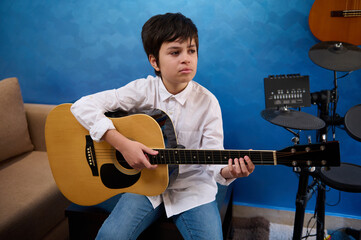 Teenager boy playing acoustic guitar at home, sitting against musical instruments and blue wall...
