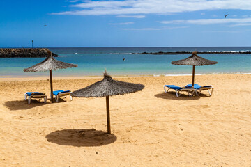 An empty beach in the off-season with straw umbrellas and blue sun lounger.  Playa del Castillo in  Caleta de Fuste, Fuerteventura, Canary Islands, Spain, - obrazy, fototapety, plakaty