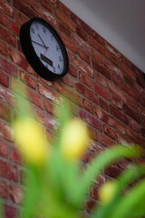 zegar okrągły biało czarny na ceglanej ścianie, z przodu rozmyty bukiet kwiatów żółto...