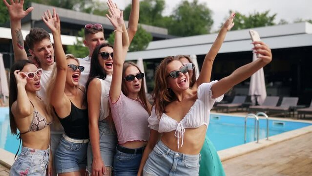 Company of female and male friends making selfie photo on phone at swimming pool in luxury beach club restaurant. Girls and boys in summer clothes posing and smiling for pov video for blog.