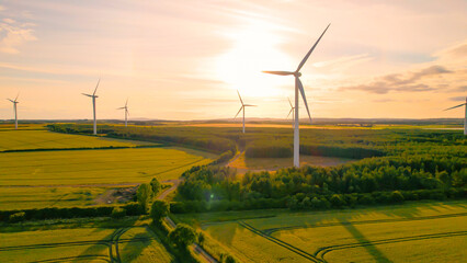 AERIAL, LENS FLARE: Colourful sunset sky above farmland and spinning windmills