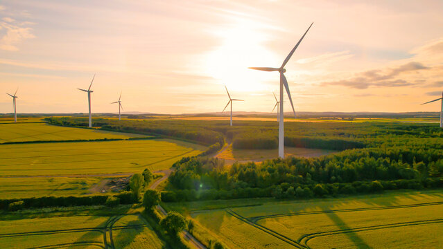 AERIAL, LENS FLARE: Colourful sunset sky above farmland and spinning windmills